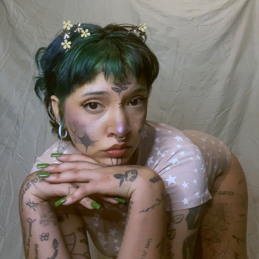 day blue poses in pink in front of a gray backdrop wearing cat ears in her short green hair.