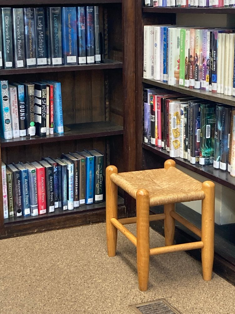 Photo shows a wooden stool sitting next to shelves of books.