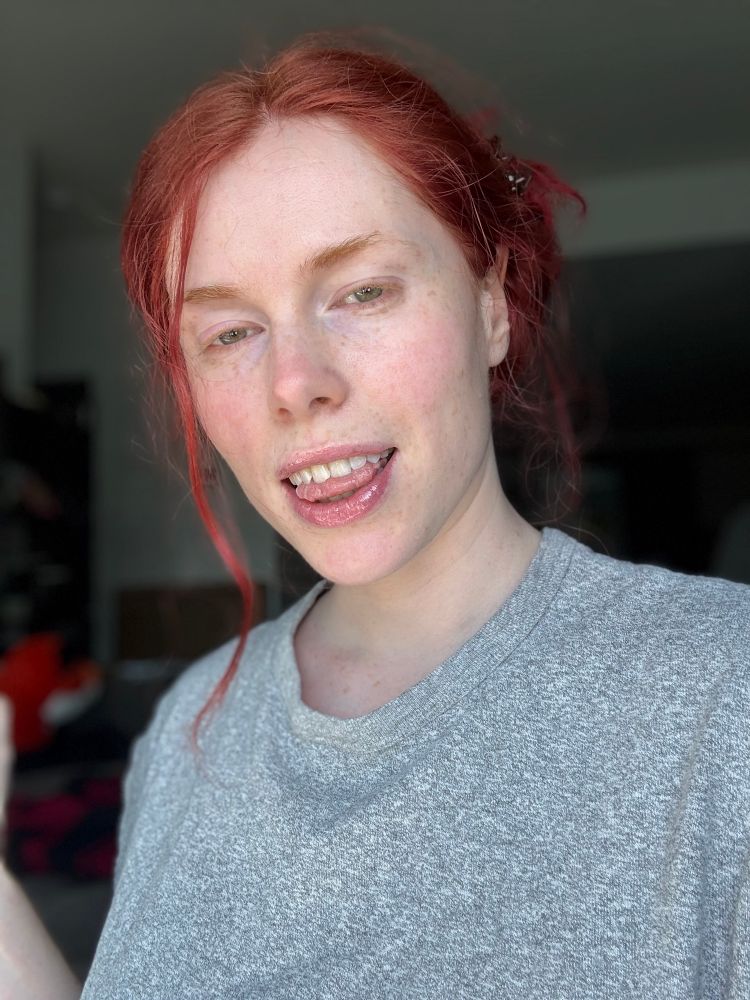 MissFox in a grey shirt, smiling with their tongue for the canners. Red hair is pulled back into a bun, stands framing their face, with a pretty blush across their cheeks.