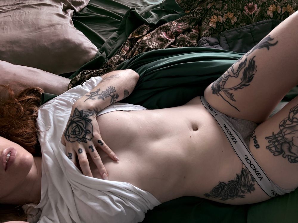 MissFox in bed, clawed hand up and lifting the front of their white shirt to tease the plump of their breasts… grey panties tugged low on their hips, framed by tattoos and soft peach bottom.