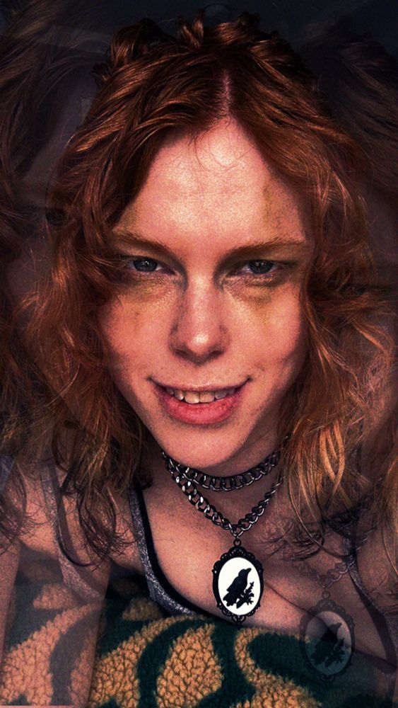MissFox looking sinister at the camera, a clown like grin on their face, gold makeup trailing like rotten clown makeup… they’ve got a cameo chain necklace on, with a black raven against a stark white background. Their hair is its usual ginger, though the tips are fox tailed, with blond and black ends.