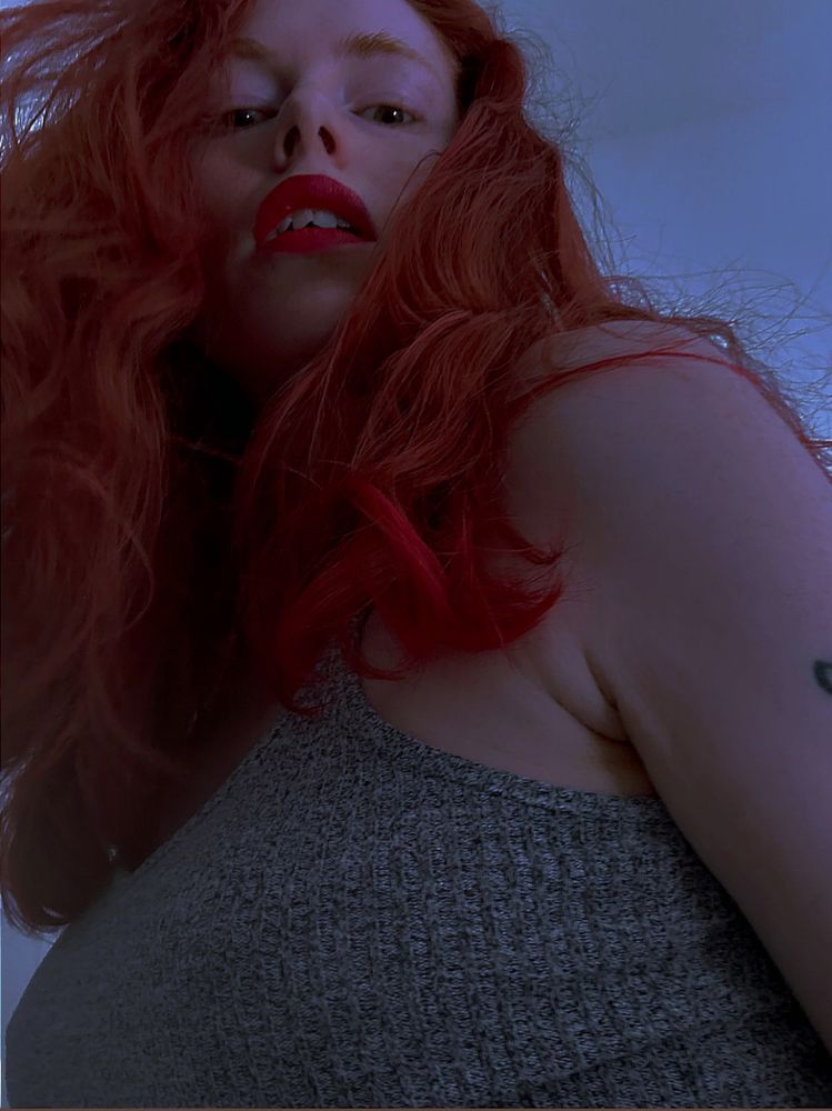 MissFox peering down, eyes glowing from within the haze of red curls. Lips ruby red as well, full and parted as they gaze at you.