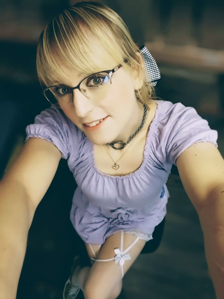 A woman sitting cross legged on a swivel chair. She is wearing a mini dress and glasses, and has a bow in her hair 