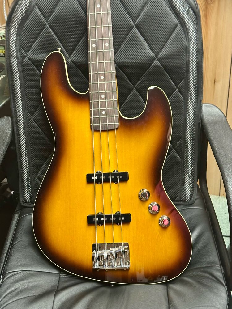 A bass guitar with a two color brown burst finish visible from the bottom of the body up to almost the 8th fret displayed on a black desk chair.