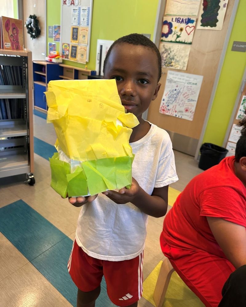 A young Black boy wearing a white t-shirt and red Adidas shorts holding a yellow and green paper sculpture in one of the library’s work rooms. 