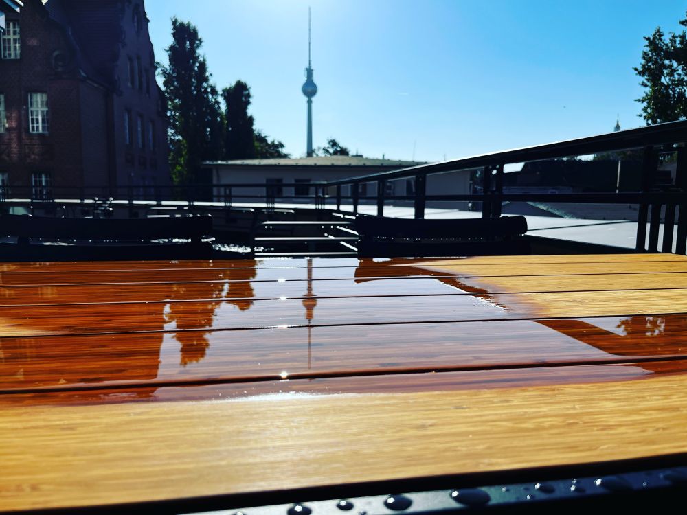 Blick Richtung Berliner Fernsehturm. 
In einer teils nassen Tischoberfläche auf einer Terrasse spiegeln sich die Konturen der Gebäude und der Turm. 