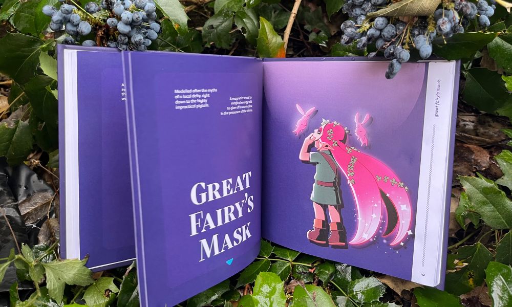 A photograph from the interior of Book of Masks, open at the Great Fairy’s Mask page. The background is purple with Link wearing the mask on the inside. The book is resting in a blueberry bush with green leaves around it