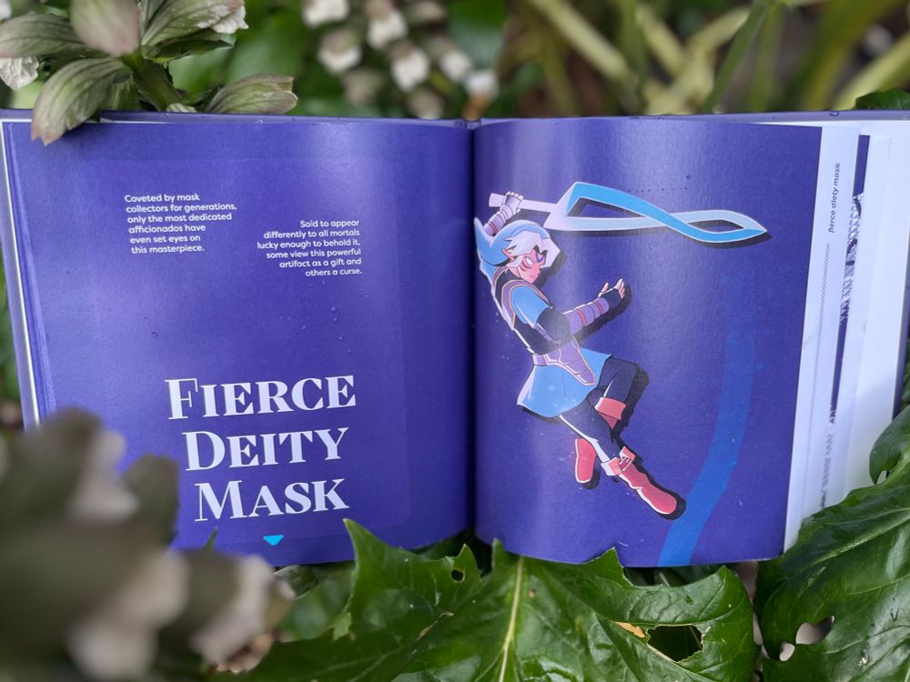 A photograph from the interior of Book of Masks, open at the Fierce Deity Mask page. The background is purple with Link wearing the mask on the inside. The book is resting amongst a patch of leaves.