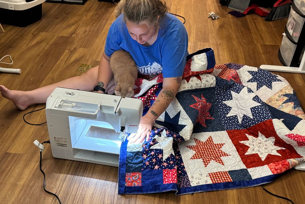 A blond woman is sitting on the floor at a sewing machine with one leg thrown out straight, working on a red white and blue quilt of eight-pointed stars on square fields of fabric, bordered by smaller blocks and a solid blue band. An orange cat is perched on her lap to assist.