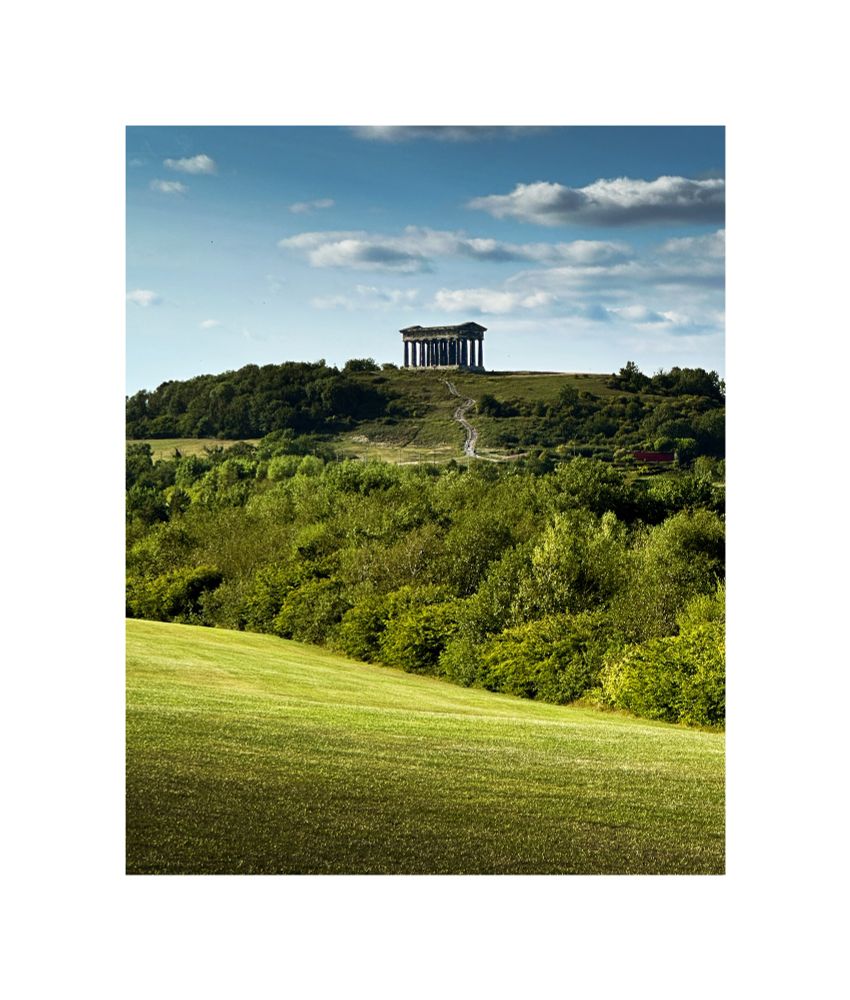 A colour image of Penshaw Monument from a distance. Rolling green hills in front and a Forrest of trees 