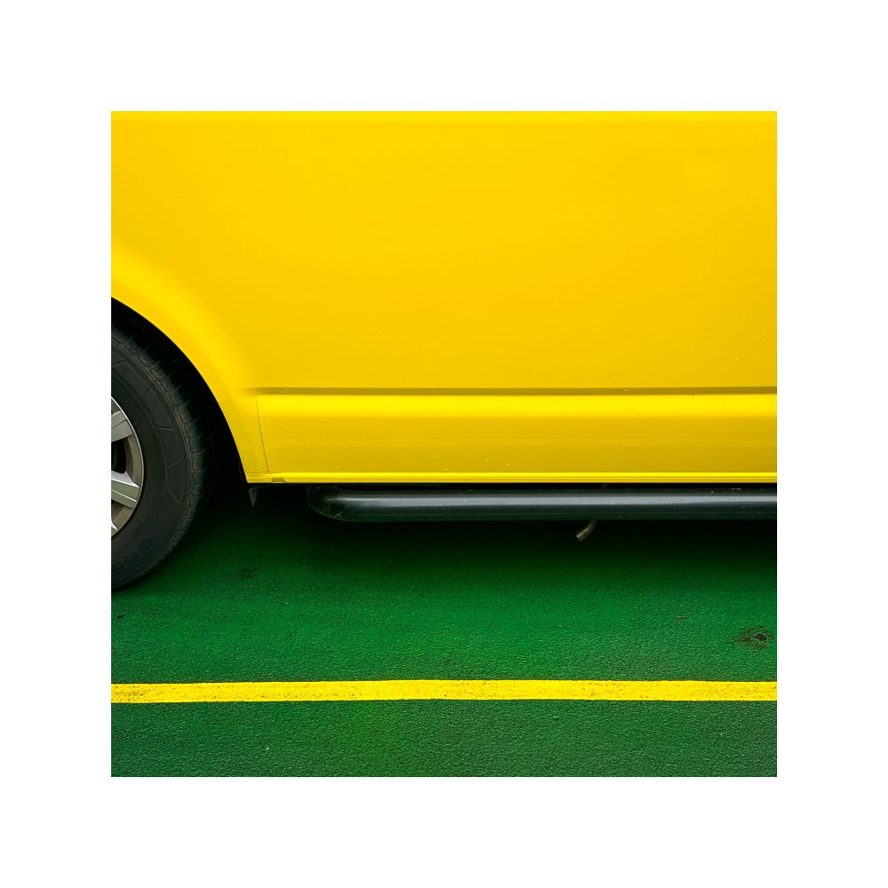 A square image of a yellow van parked on the green floor of the ferry with a yellow strip across the bottom of the image. You can only see the bottom half of the van with part of the wheel on the left