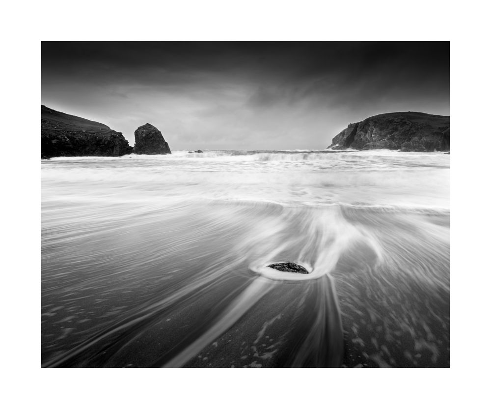 Black and white image of a rock in the foreground with water movement around it leading to the sea. Each side is the dark cliffs and rough waves are in the middle 