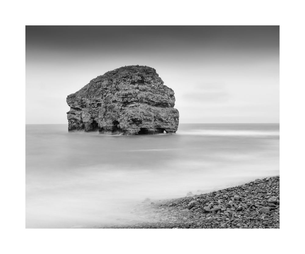 A black and white seascape with a large rove surrounded by calm milky water
