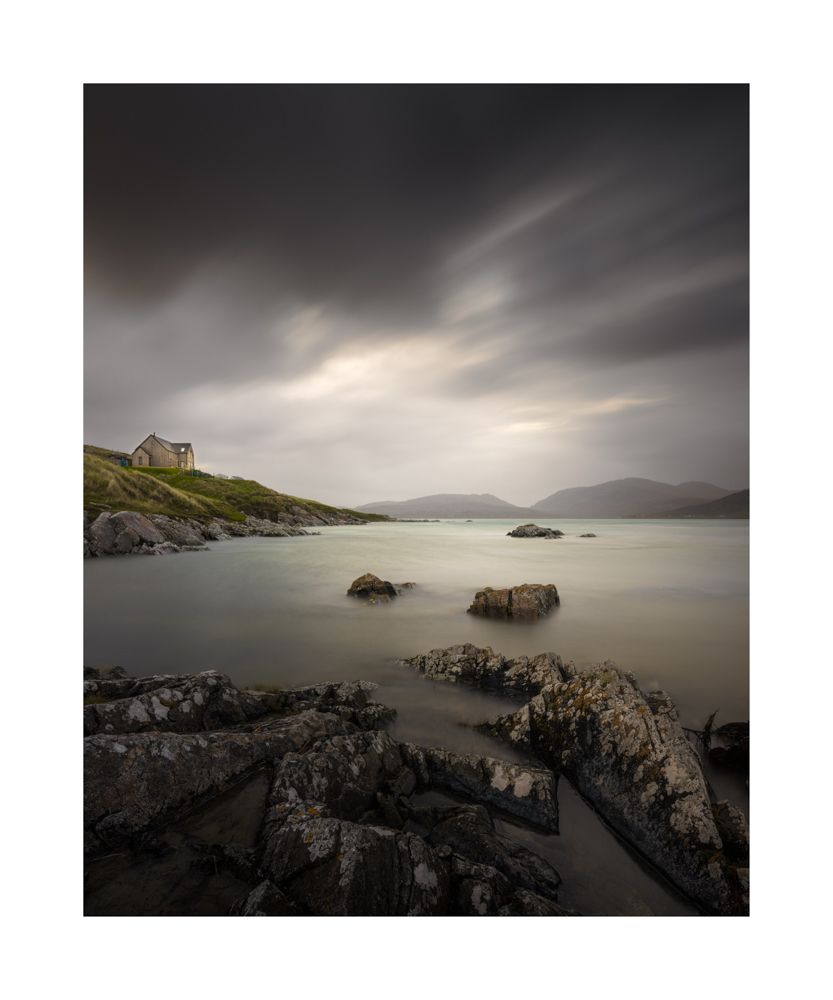 Colour image of the water at Luskintyre . Rocks in the foreground a house to the left and plenty movement in the clouds 