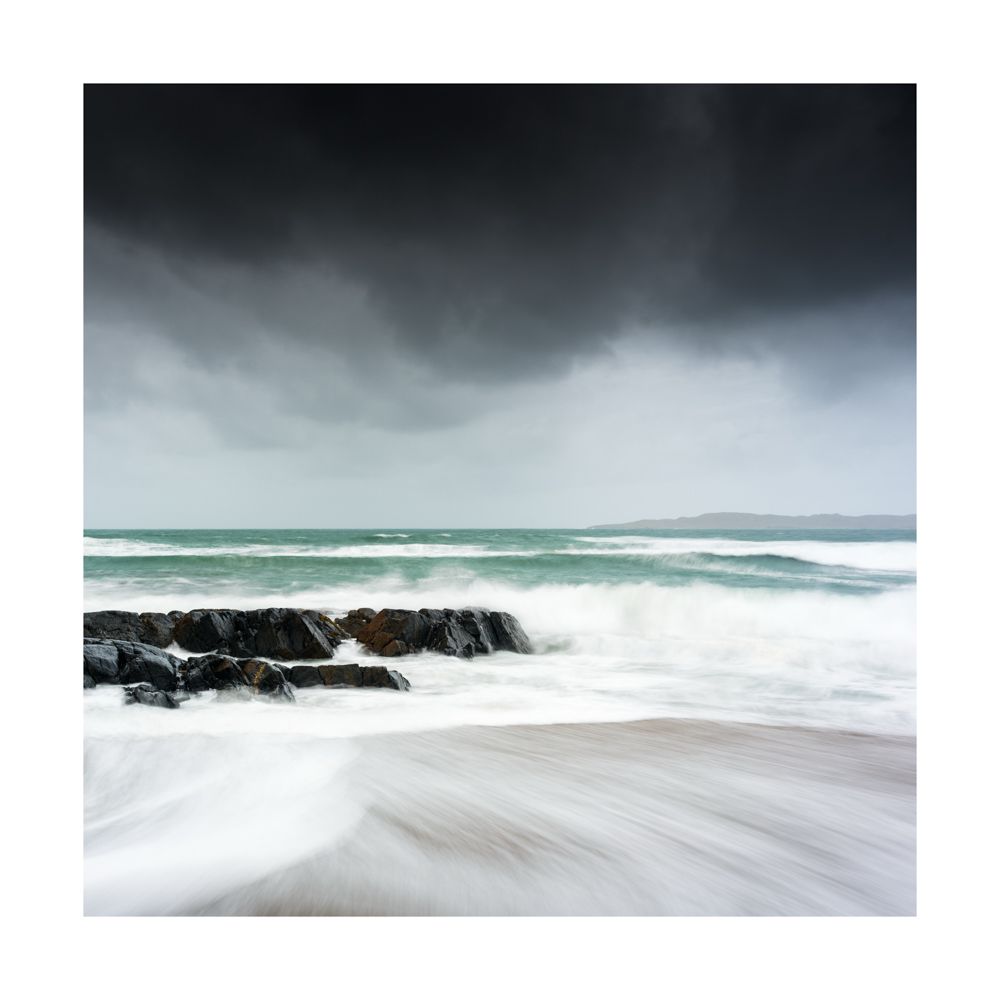 Rocks jutting in from the left surrounded by water crashing up against them, demonstrating the energy of the sea. The water is turquoise and there are dark clouds above