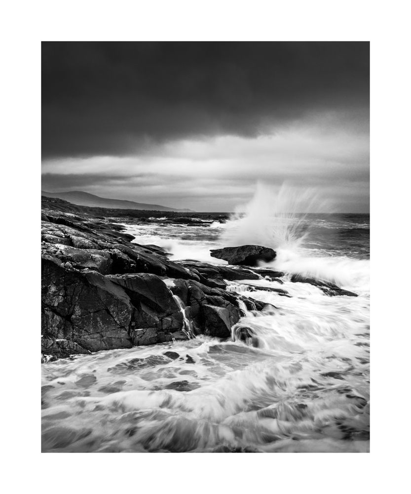 Black and white seascape. Rocks to the left with waves smashing against them 