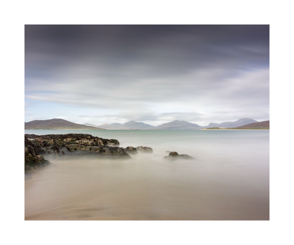 Colour image of a beach with a small bit of sand then the turquoise water. Rocks coming in from the left and mountains in the background 