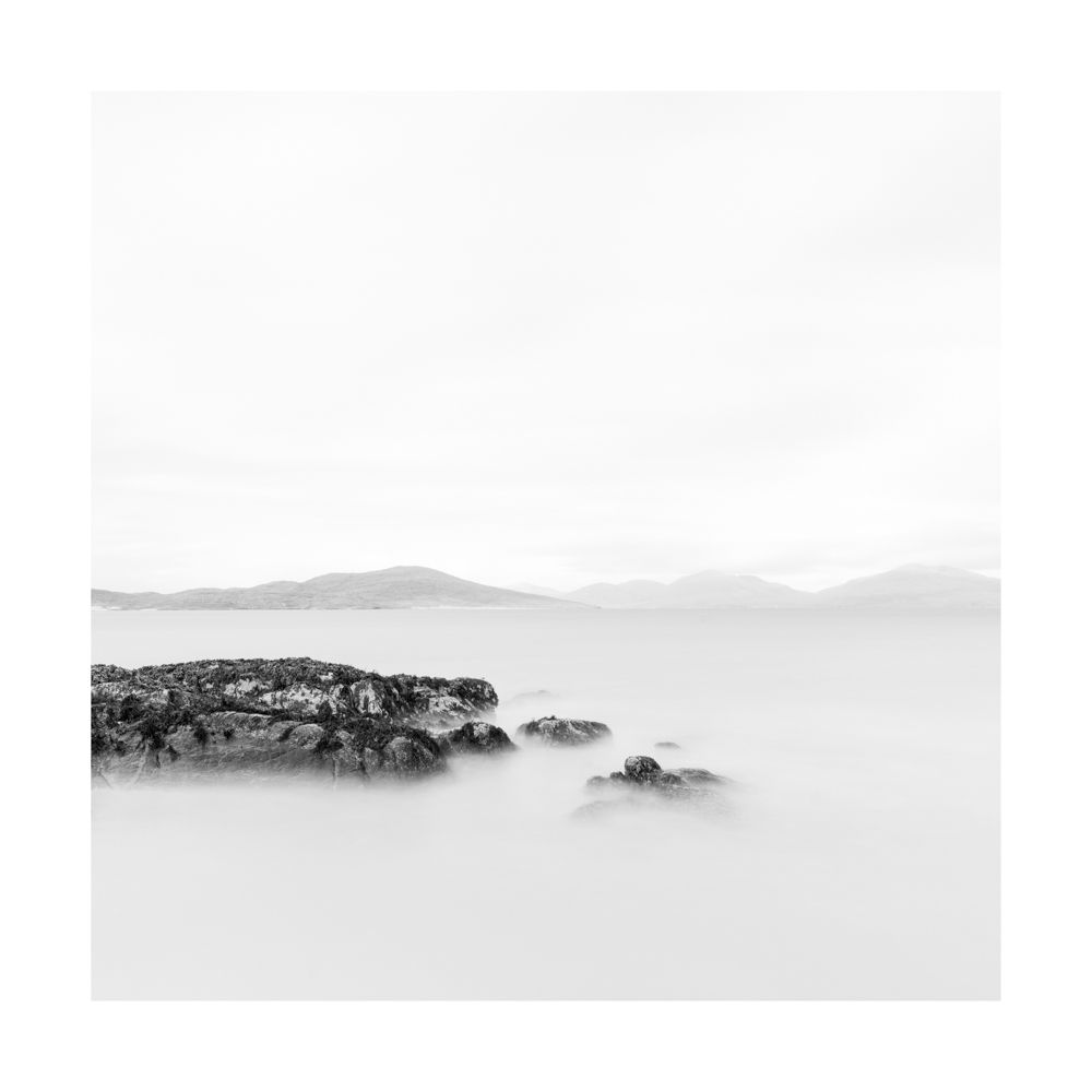 Square black and white image of a dark rock jutting out from the left surrounded by calm smooth water with hills in the distance 