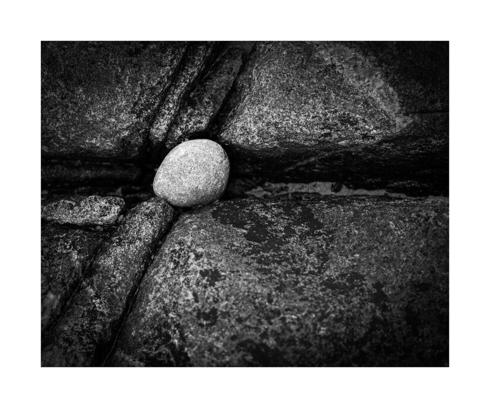 Black and white image of a white round rock stuck in a crevice on other rock