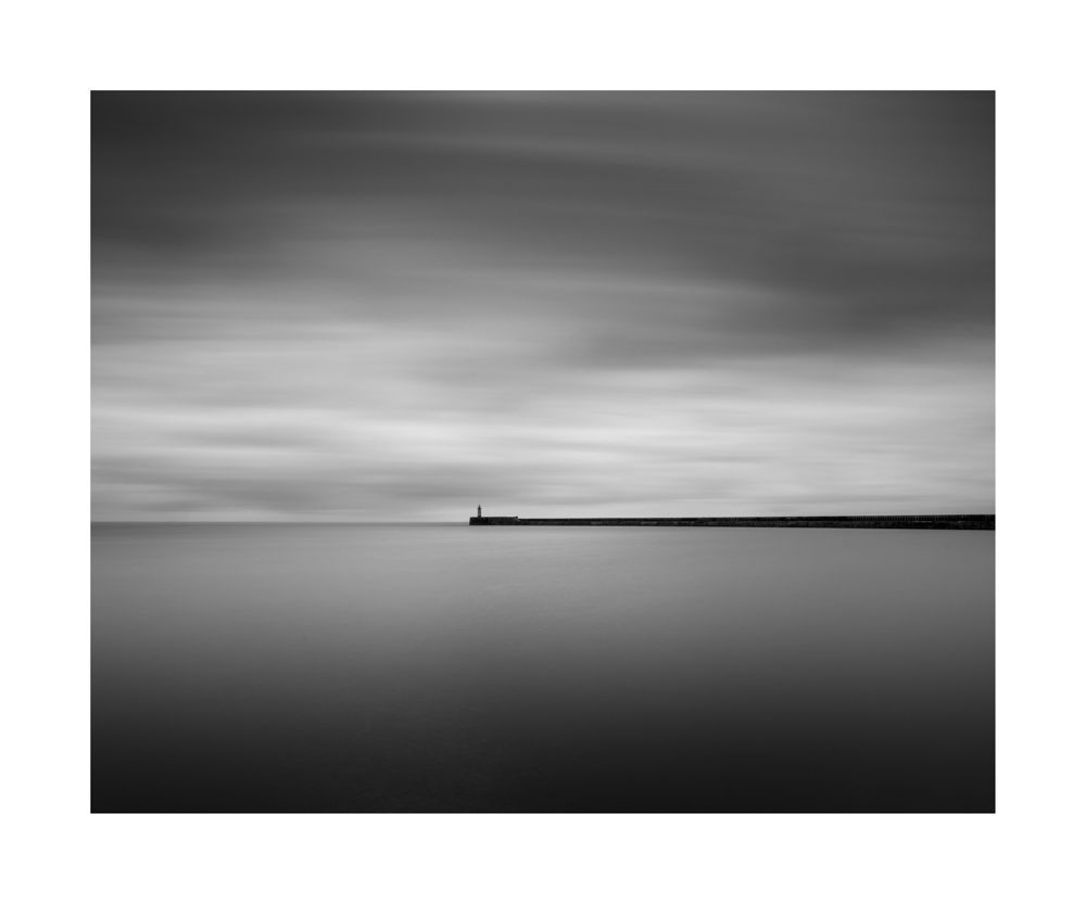 Black and white seascape. Lots of calm water and sky with a pier and lighthouse gutting in from the right side