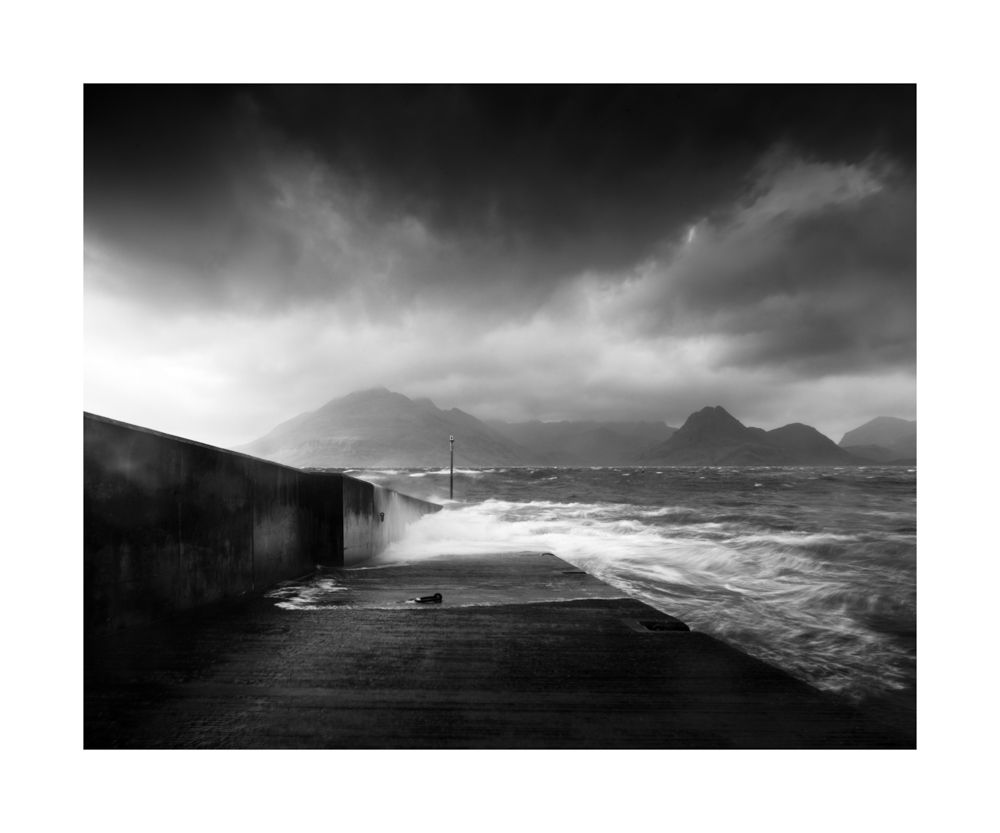 A dramatic black and white image of the pier heading straight out to see. Waves smashing against the harbour wall with mountains in the background