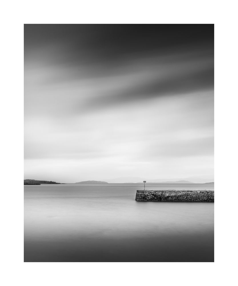 Black and white image of a pier jutting out from the right. The water is calm and clouds show movement in the sky
