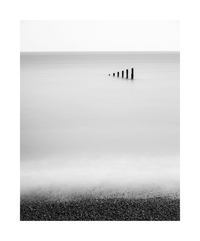 A simple black and white image of the sea. With a small bit of the beach then calm water surrounding the 6 groynes appearing out of the water 