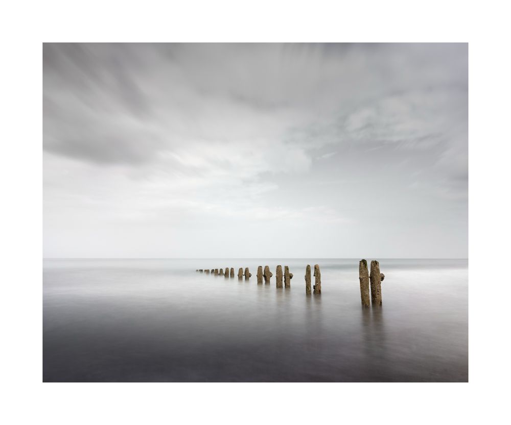 A row of groynes heading out to sea surrounded by calm milky water 