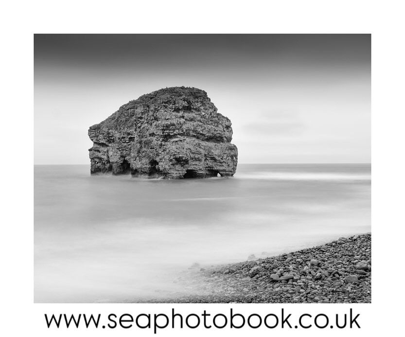 A black and white image of Marsden Rock in the northeast coast of the UK surrounded by calm water with a little bit of the rocky shoreline shown in the bottom right corner 