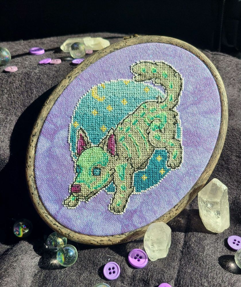 A finished cross stitch on purple fabric, displayed in a hoop with some decorations scattered about. The subject of the stitch is a playful grey wolf-dog with glowing bones above the fur. They stand in front of a circular gradient sky background.