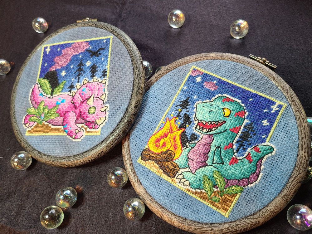 Two hooped cross stitch pieces of dinosaur plushies. They both have a night scene background. The left is a pink triceratops with blue spots, laying down with its legs splooted. The right is a teal tyrannosaurus with red stripes and a purple belly. There is a campfire in front of the trex.