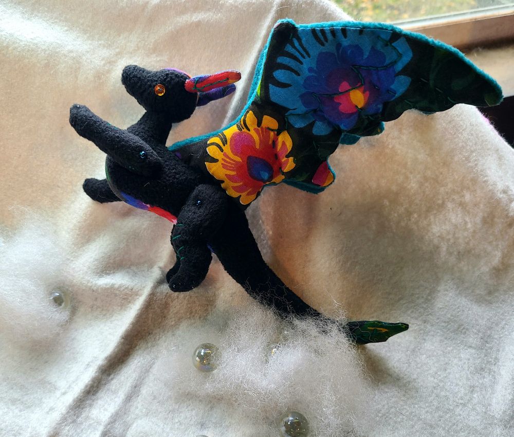 A black dragon-like plush with floral fabric accents on the belly and wings. It appears to be 'flying' through some cotton clouds.