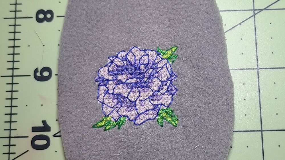 A light purple/blue flower with small green leaves cross stitched onto a piece of grey fleece.