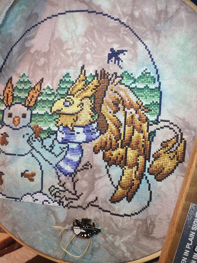 A partially finished cross stitch of a brown/gold gryphon building a snowman.