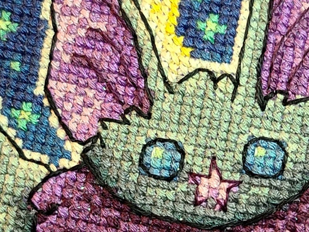A close up of a cross stitch depicting a grey bat with purple inner ears and nose, and blue eyes.