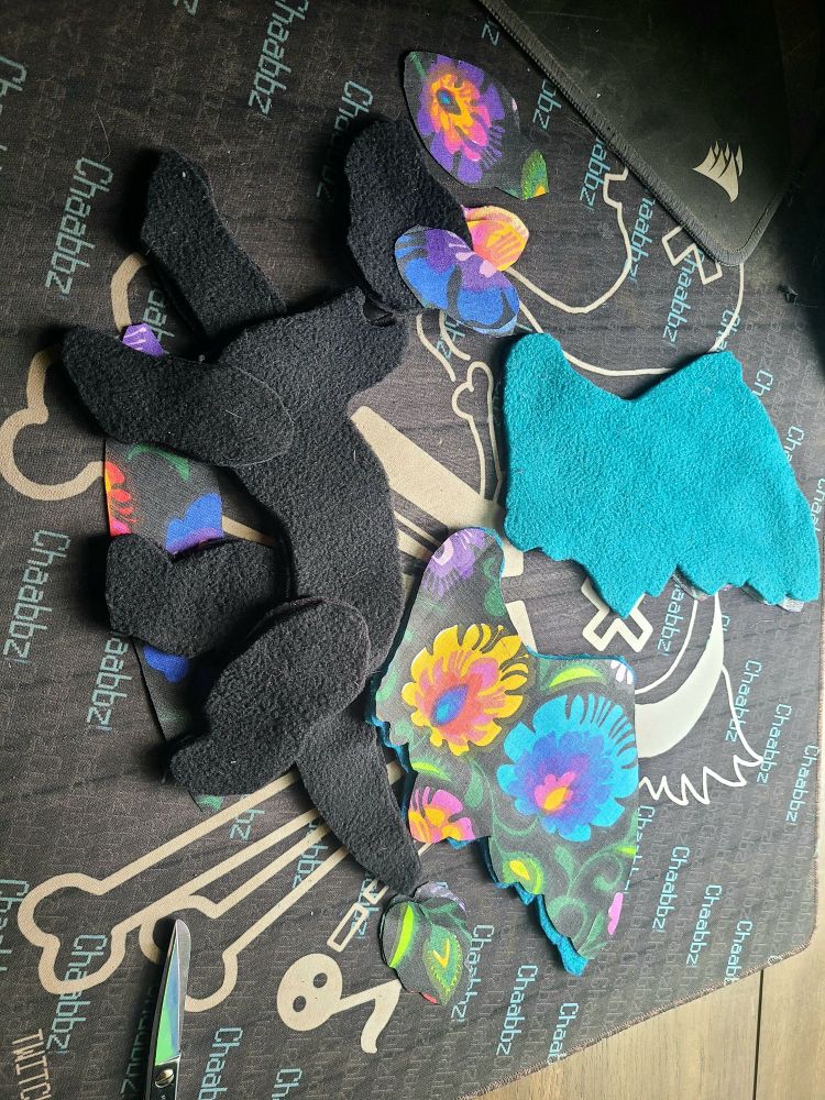 Pieces of a dragon plushie cut out and laid on a surface ready to be sewn up. The fabrics are black and teal fleece, and a flower printed cotton.