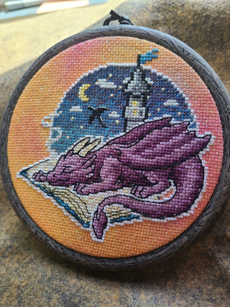 A cross stitch on bright orange/pink sparkle fabric. It shows a purple dragon sleeping on an open book. The background is a lone tower in the night sky, in which you can see the silhouette of a flying dragon.