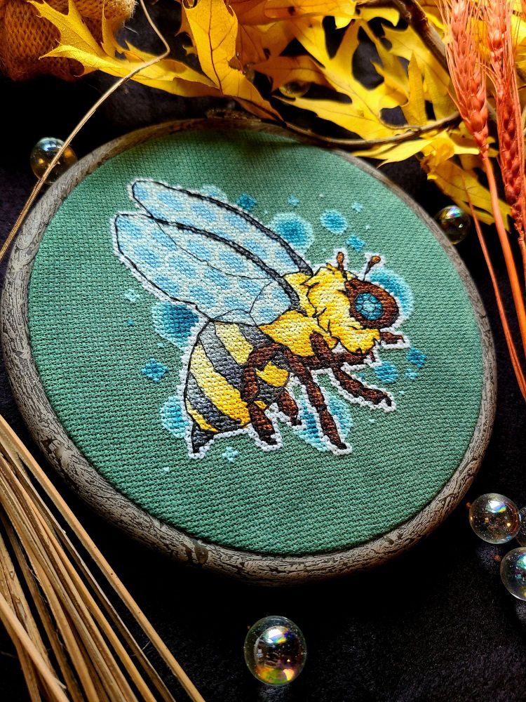 A hooped cross stitch of a brightly colored bee on green fabric. There are blue bubbles serving as a background for the bee. The hoop is surrounded by leaves and marbles.