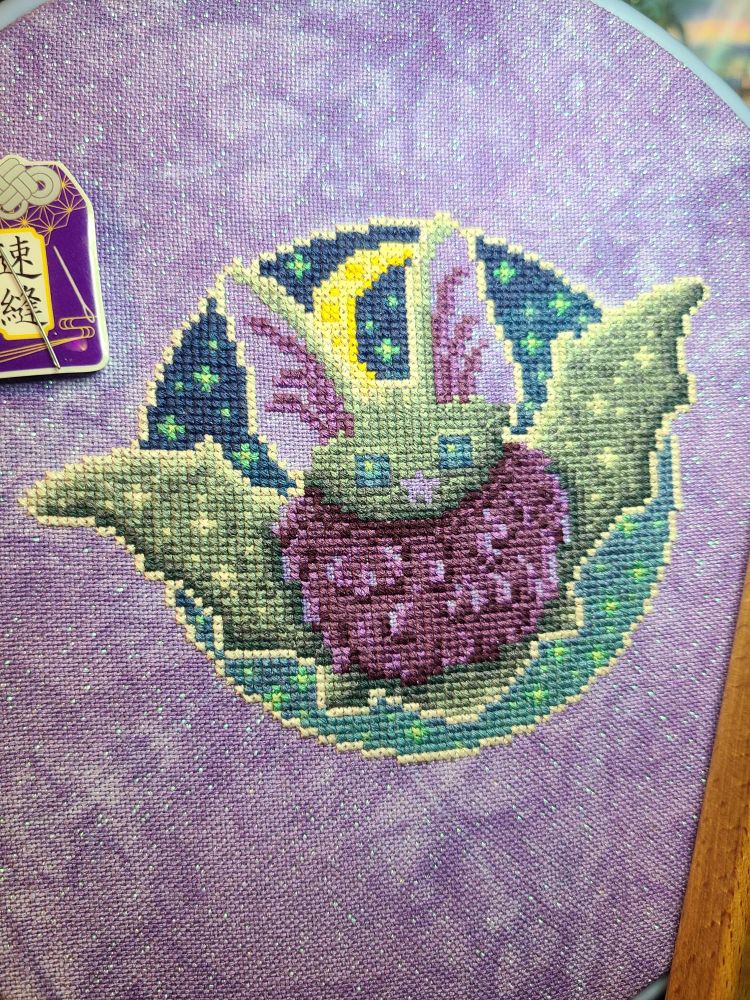 A nearly complete cross stitch of a grey bat with purple fur and inner ears with a starry gradient sky. The fabric is purple and sparkly.