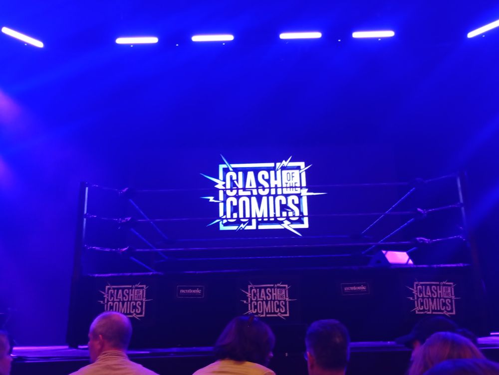A lot stage, on which a neon sign reads "Clash of the Comics". 