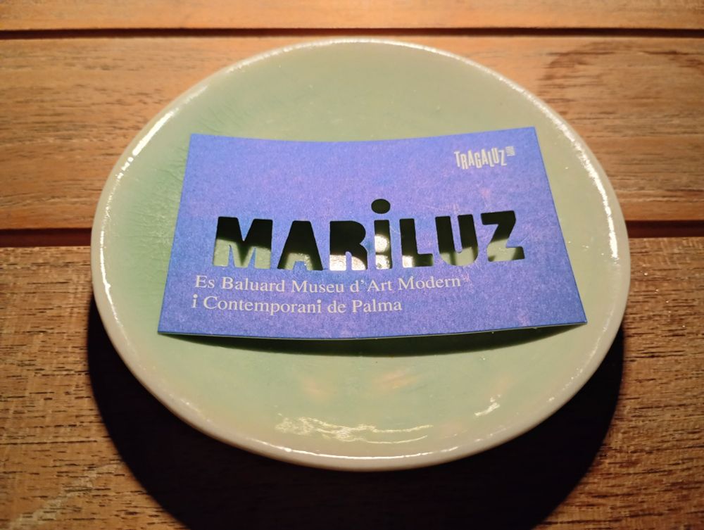 Business card for the bar, which has ",MARiLUZ" cut out, lying on a saucer on a wooden table. 