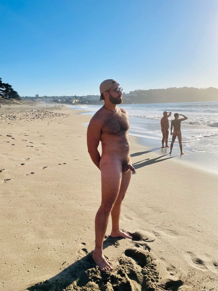A guy stands naked on a beach in the sun with two guys in the distance.  He his tall hairy and beaded and wearing a hat 