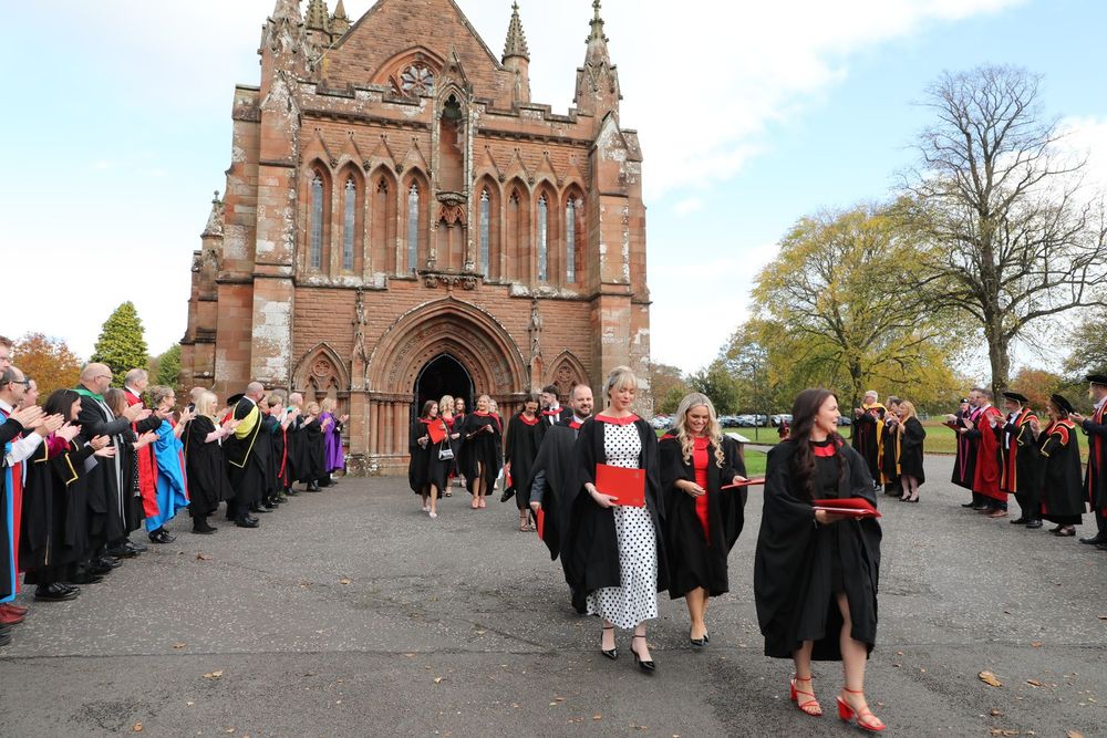 Graduates in academic robes and sashes walk away from a historic church, celebrating their commencement ceremony.