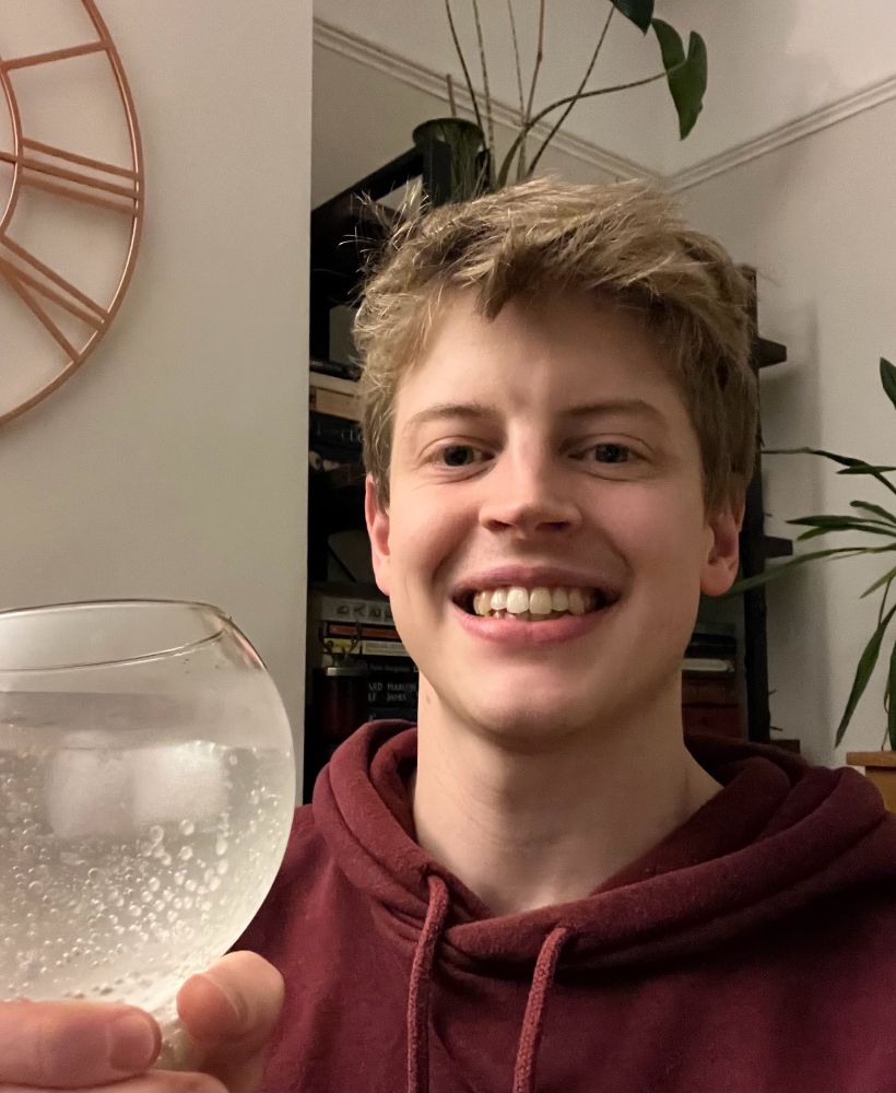 Selfie of Sam wearing a dark red hoodie and holding a glass filled with clear liquid, bubbles and ice