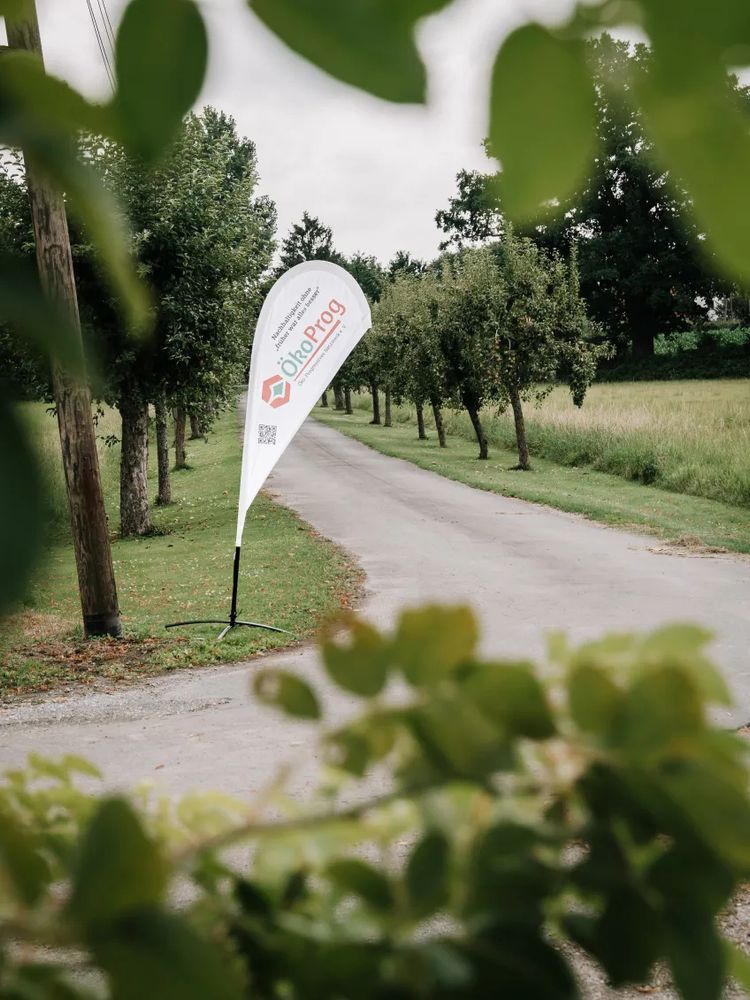 Beachflag mit dem ÖkoProg-Logo in der Einfahrt zur Event Location, einem Weg gesäumt von Obstbäumen.