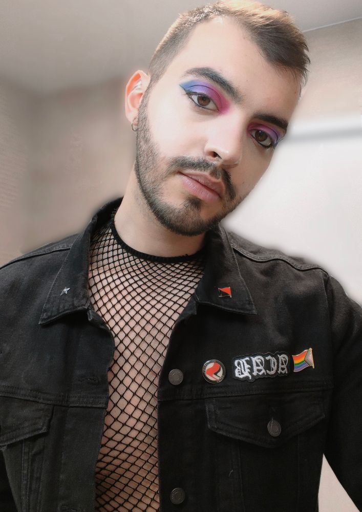 Selfie de moi maquillée avec un fard à paupières dégradé aux couleurs du drapeau bisexuel, bleu violet rose
J'ai un fishnet sur ma poitrine nue, et une veste en jean noir avec quelques pin's et patch : les symboles antifa, LGBTQIA+ etc