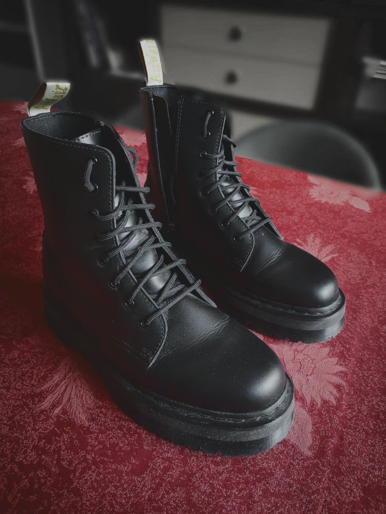 Mes Dr Martens vegan d'occasion, posées sur ma table. Elles sont monochromes noires, et n'ont pas le traditionnel fil jaune autour de la base de la chaussure
Elles ont une semelle assez épaise (~5 cm) et leurs étiquettes à l'arrière de la cheville sont blanches 