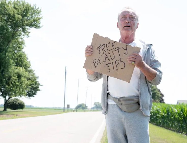 Udo Kier as Mr. Pat hitchiking in SWAN SONG (Todd Stephens, USA, 2021) holding a sign that says "FREE BEAUTY TIPS"