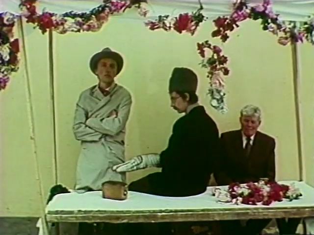 L'homme, au stand de la brocante, tentant de vendre sa bombe aux côté d'un homme portant un poisson en peluche. Des fleurs sont accrochées au stand.