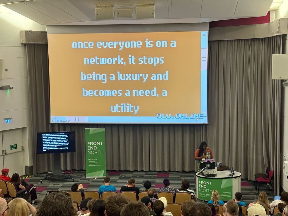 Olu presenting a slide that reads "once everyone is on a network, it stops being a luxury and becomes a need, a utility"